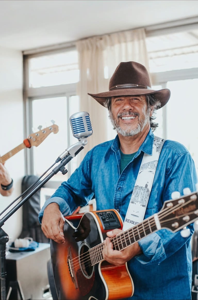 Gleno Rossi de chapéu marrom e camisa jeans, sorrindo com o violão ao lado do microfone